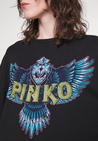 Persona che indossa una maglietta nera con una grande grafica di un uccello alato blu e il testo giallo-verde "PINKO" sul petto, con capelli castani ricci in vista.