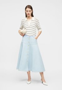 Γυναίκα που φοράει ένα κρεμ ριγέ πλεκτό τοπ, φούστα midi σε ανοιχτό μπλε με κουμπιά, λευκές ψηλές γόβες και κρατάει μια κρεμ τσάντα χειρός.