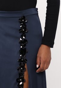 Jūras zila satīna svārku ar sānu šķēlumu un melnu ziedu aplikāciju kolonnu ar spīdīgām pērlēm. Gluda tekstūra un pieguļoša forma.