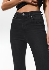 Mustat korkeavyötäröiset denim-farkut, joissa on sileä pinta, viisi-taskuinen muotoilu, metallinen nappikiinnitys ja hienovaraiset ompeluyksityiskohdat.