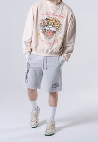 Sudadera beige con gráfico de tigre y letras; pantalones cortos grises con detalles negros; calcetines blancos; zapatillas beige con suela verde.