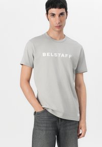 Γκρίζα βαμβακερή μπλούζα T-shirt με στρογγυλή λαιμόκοψη, κοντά μανίκια και άσπρο κείμενο "BELSTAFF" στη διάσπαση του στήθους. Συνδυάζεται με σκούρες γκρίζες τζην.