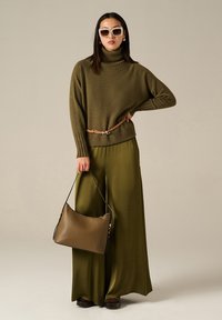 Maglione oversize verde oliva con colletto alto, cintura beige e pantaloni larghi verde oliva. Accessoriato con una borsa marrone e occhiali da sole bianchi.