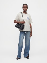 Femme portant une chemise beige à manches courtes, un jean bleu à jambes larges avec des fentes à l'avant, des chaussures noires à bouts pointus et un sac à bandoulière noir.