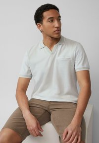 Lichtgrijze poloshirt met V-hals, korte mouwen en een borstzak. Heeft subtiele contrasterende biezen op de mouwen en kraag. Bruine shorts.
