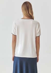 White short-sleeve top with a rounded neckline, featuring black piping along the edges. The back is smooth with a relaxed fit.