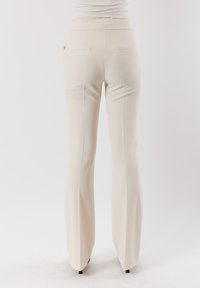 Pantaloni beige a campana con vita aderente, due tasche posteriori e un tessuto liscio, che si estende fino al pavimento coprendo delle scarpe nere.
