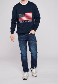 Sudadera azul marina con un diseño de bandera de EE. UU. en rojo y blanco, con el texto "U.S. POLO ASSN." debajo. Combinada con jeans azules y zapatillas grises.