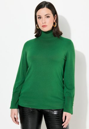 Grüner Rollkragenpullover aus einem leichten, strukturierten Strick. Lange Ärmel mit Knopfdetails an den Bündchen. Kombiniert mit glänzenden schwarzen Hosen.