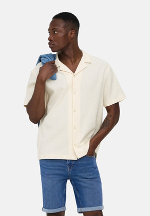 Homme portant une chemise crème à manches courtes et un short en jean retroussé, tenant une veste bleue pliée sur l'épaule, regardant vers sa gauche.