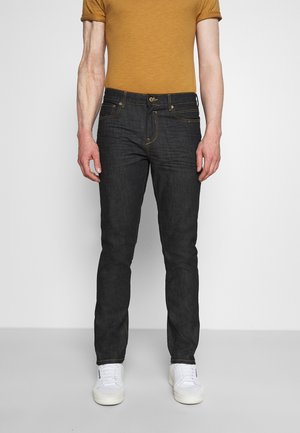 Homme portant un jean slim noir, une chemise manches courtes moutarde et des baskets blanches, debout devant un fond clair uni.