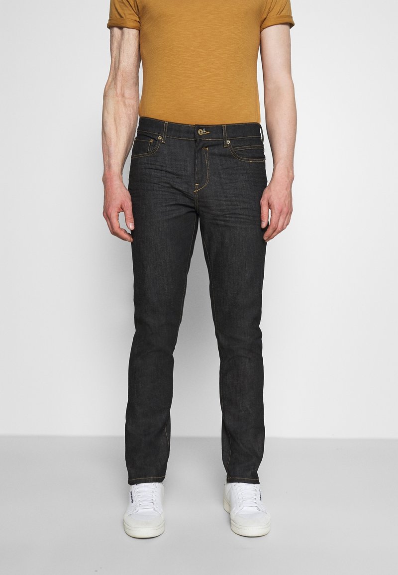 Homme portant un jean slim noir, une chemise manches courtes moutarde et des baskets blanches, debout devant un fond clair uni.