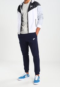 Chaqueta de Nike en blanco con detalles en negro, camiseta gris, pantalones de chándal azul marino y zapatillas azules. Presenta un diseño deportivo y una silueta ajustada.