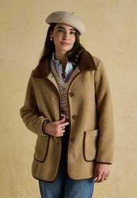 Beež fliiskatte kätki pruunist korduroost kraega, kahe esifrakta ja esi nuppudega, mis on kontrastse äärega. Mudel kannab nende all kihilisi rõivaid.