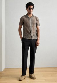 Cămașă cu mânecă scurtă, cu un model în tonuri de maro, asortată cu pantaloni negri. Modelul poartă ochelari de soare și pantofi bej. Material neted, texturat.