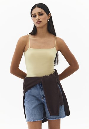 Giovane donna con lunghi capelli scuri, indossa un top color beige, pantaloncini di jeans e un maglione scuro annodato in vita, in piedi con le mani dietro la schiena.