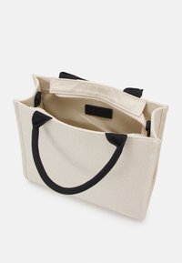 Beige lærredstaske med sorte stofhåndtag, lynlåslukning og en indvendig lomme. Har en struktureret rektangulær form.
