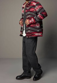 Veste en mélange de laine avec des motifs géométriques rouges, gris et blancs, superposée à une chemise grise, associée à un jean foncé et des chaussures noires.