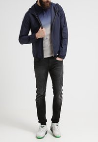 Marinblå huva-jacka, grå gradient polo, mörka jeans och grå sneakers med gröna accenter. Texturerad tyg, elastiska ärmslut och dragkedjestängning.