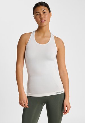 Frau trägt ein weißes ärmelloses, figurbetontes Tanktop und grüne Sportleggings, steht mit neutralem Gesichtsausdruck vor einem einfarbigen Hintergrund.