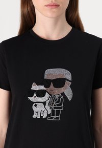 Svart bomull T-skjorte med rhinestones i designet av en figur med solbriller ved siden av en stilisert hvit katt, begge med detaljerte aksenter.