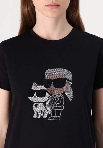 T-shirt en coton noir ornée d'un design en strass représentant un personnage porte des lunettes de soleil aux côtés d'un chat stylisé blanc, tous deux avec des accents détaillés.