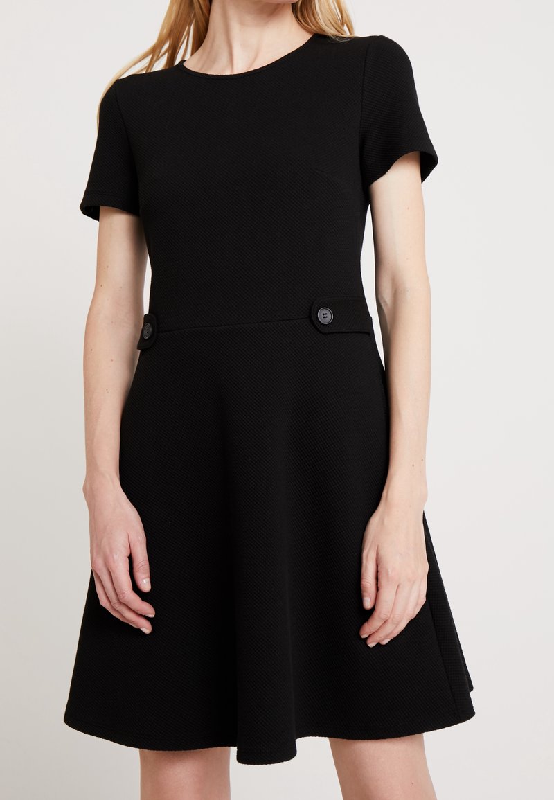 Femme portant une robe noire à manches courtes en ligne A avec un tissu texturé et deux boutons décoratifs à la taille.