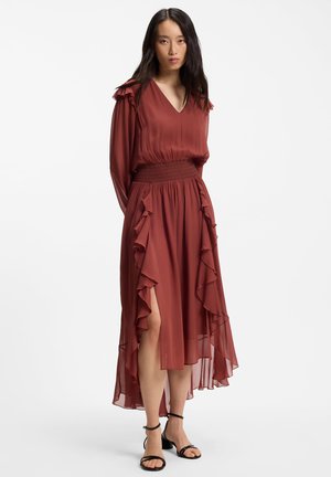 Femme debout portant une robe à manches longues couleur rouille avec des volants, un ourlet asymétrique, une fente sur le côté et des sandales à talon carré noires avec bride à la cheville.