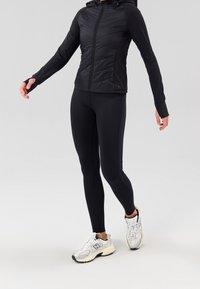 Chaqueta acolchada negra con capucha, mangas largas y orificios para los pulgares; combinada con leggings ajustados negros y zapatillas deportivas gris claro con detalles en negro.