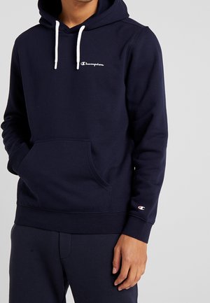 Homme portant un sweat à capuche Champion bleu marine avec cordons blancs, poche avant et logo sur la poitrine et la manche sur un fond gris.