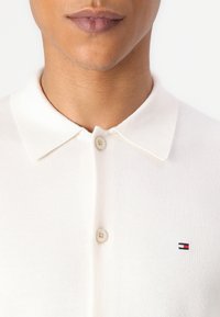 Biała koszulka polo o miękkiej fakturze, z klasycznym kołnierzykiem i trzema guzikami. Małe haftowane logo w kolorach granatowym, czerwonym i białym na piersi.