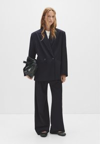 Blazer bleumarin cu dungi fine, cu două rânduri de nasturi, pantaloni largi asortați și o geantă neagră lucioasă. Ținuta are un croi lejer și o textură netedă.