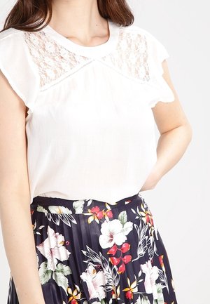 Witte mouwloze top met kantdetails op de schouders en halslijn, gecombineerd met een bloemenrok met plooien in het donker, versierd met levendige bloesems.