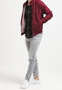 Chaqueta bomber roja con forro de cuadros sobre una camiseta negra, combinada con jeans gris claro y zapatillas blancas. Patrón geométrico sencillo en la camiseta.