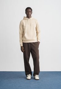 Béžová mikina s predným vreckom, v kombinácii s tmavohnedými teplákmi s bielym logom. Model má na nohách biele tenisky a stojí na modrej podlahe.