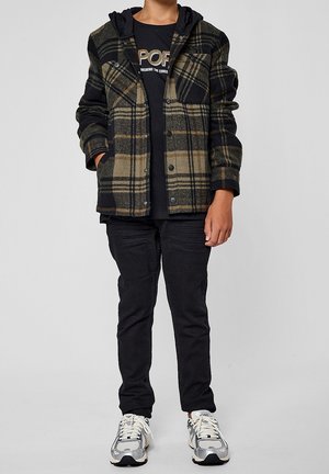 Osoba ubrana w czarno-oliwkową kraciastą kurtkę zapinaną na guziki, czarną bluzę z kapturem, czarne spodnie i białe trampki, stojąca z rękami w kieszeniach.