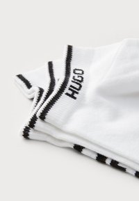Hvide sokker med sorte striber og "HUGO" tekst. Blødt stof med ribbet tekstur. Viser tre stakkede par, der viser detaljen af designet.