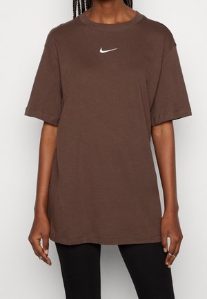 Personne portant un t-shirt Nike marron ample à manches courtes avec un logo swoosh blanc Nike sur la poitrine, associé à un pantalon noir.