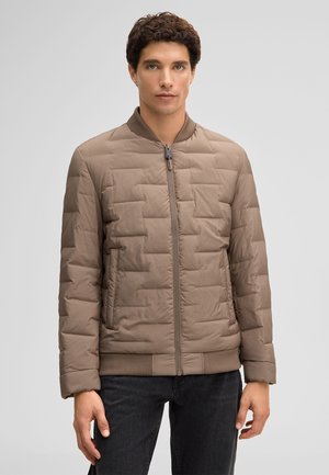 Mężczyzna z krótkimi kręconymi włosami, ubrany w jasnobrązową pikowaną kurtkę na zamek i ciemne spodnie, stoi na tle jednolitego szarego tła.