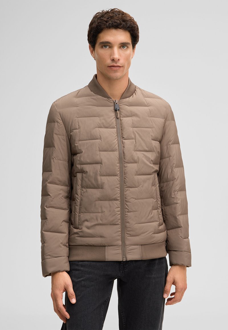 Uomo con capelli corti e ricci che indossa una giacca trapuntata marrone chiaro con cerniera e pantaloni scuri, in piedi davanti a uno sfondo grigio semplice.