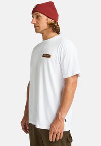 Hvid kortærmet t-shirt med rund hals, der har et brunt rektangulært logo-patch på brystet. Komplementeres med olivengrønne bukser.