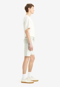 Camisa blanca de algodón de manga corta, pantalones cortos de mezclilla claros, calcetines blancos y zapatillas crema con suelas marrones, mostrados de lado sobre un fondo liso.