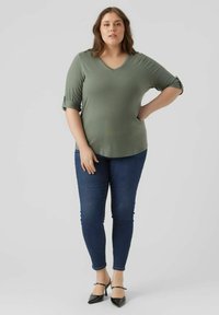 Olijfgroene V-hals shirt met driekwart mouwen, gecombineerd met donkerblauwe skinny jeans en zwarte hakken. Glad materiaal, losse pasvorm.