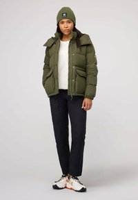 Olivgrüne Steppjacke mit hohem Kragen, Fronttaschen und Reißverschluss, kombiniert mit einer passenden Mütze und schwarzen Hosen. Weiße Sneakers sind ebenfalls enthalten.