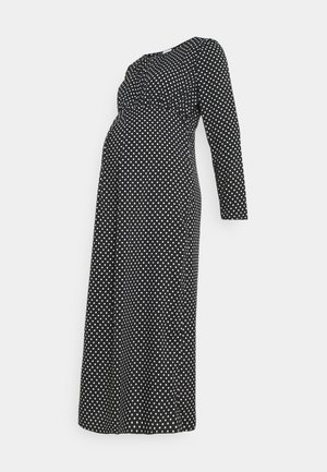 Robe de maternité noire à manches longues avec des pois blancs et une taille ajustée sur fond blanc.