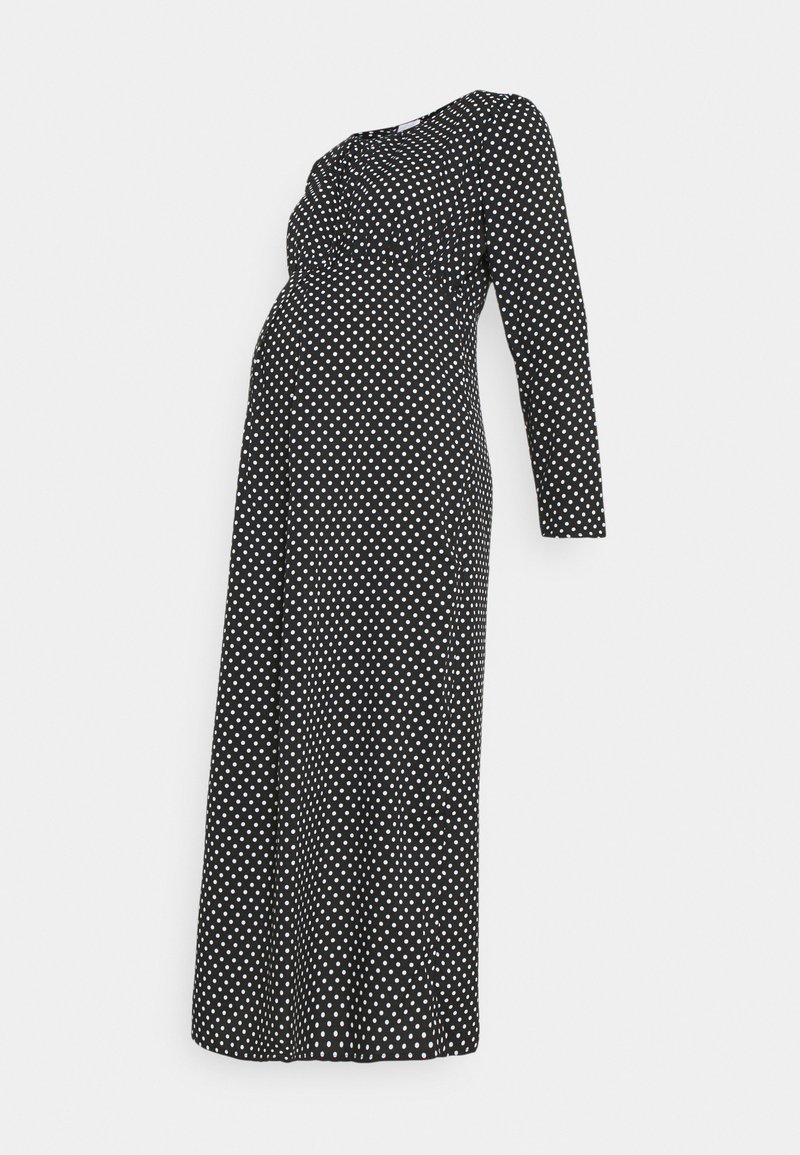 Robe de maternité noire à manches longues avec des pois blancs et une taille ajustée sur fond blanc.