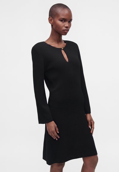Femme portant une robe noire à manches longues avec un petit décolleté en forme de trou de serrure fermé par un seul bouton doré.