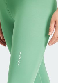 Leggings compressivi verdi realizzati in tessuto liscio, con un logo bianco nella parte bassa e cuciture piatte per ridurre l'attrito.