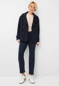 Marineblauwe dubbelgeknoopte zeemanjas met grote knopen, crèmekleurige sweater, donkere spijkerbroek en witte sneakers, tegen een effen achtergrond.