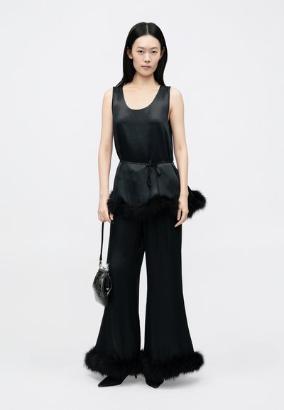 Top negru din satin, fără mâneci, cu talie ajustabilă prin șnur, asortat cu pantaloni evazați negri, cu margini din blană la tiv. Model ținând o geantă neagră.